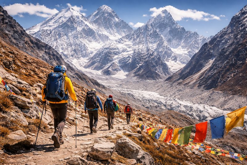 vandrare-mot-mount-everest-base-camp-himalaya-16×9 Vandrare på trekkingled mot Mount Everest Base Camp i Himalaya, med snötäckta bergstoppar, glaciärdal och färgglada böneflaggor i förgrunden.